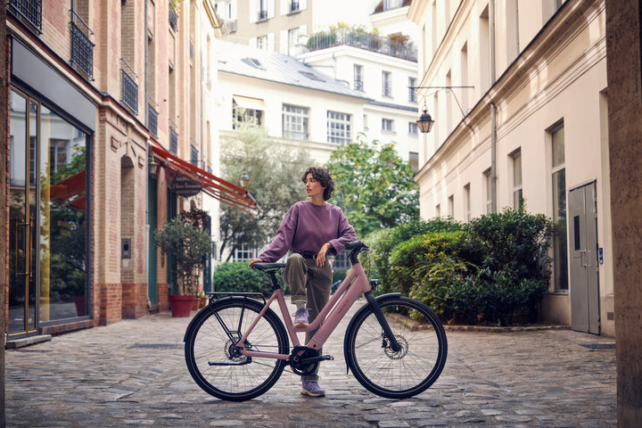 Young Woman on Cobblestone Street Standing WIth Riese & Muller Culture Mixte E-Bike in Colour Blossom