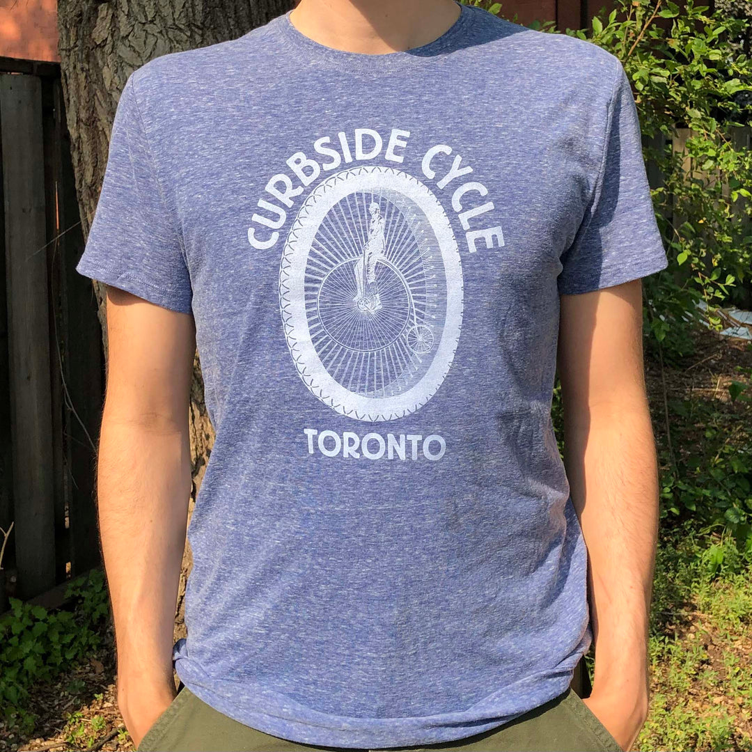 Curbside Cycle Old School Logo T-Shirt with Bike Wheel in Colour Blue on Male Model Outside in the Sun